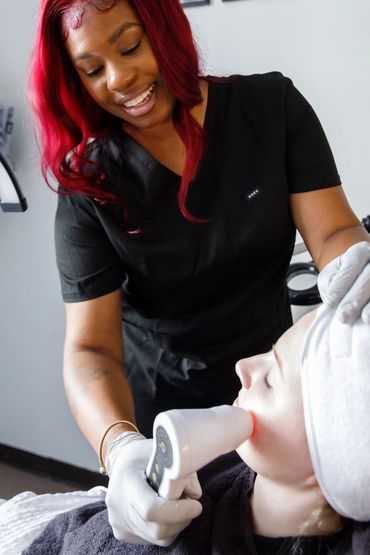 A smiling beautician performs a facial treatment on a client using a handheld device.