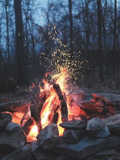 A glowing campfire with sparks flying in a forest at dusk.