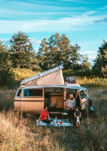 Family camping with a van in a sunny, grassy field surrounded by trees.