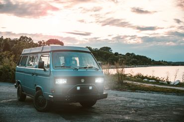 Vintage blue Volkswagen van parked near a lake during sunset with headlights on.