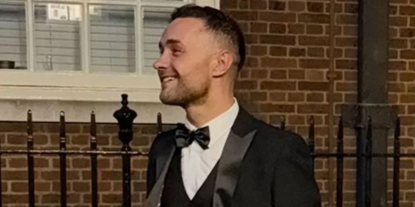 Man in tuxedo smiling outdoors near brick building.