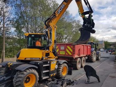 Yellow JCB excavator loading debris into red trailer on a street.