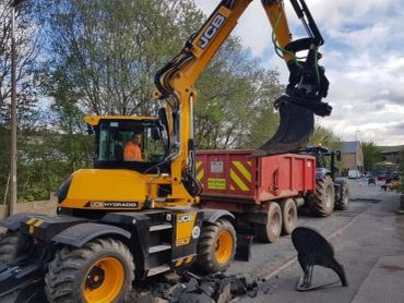 Yellow JCB excavator loading debris into red trailer on a street.