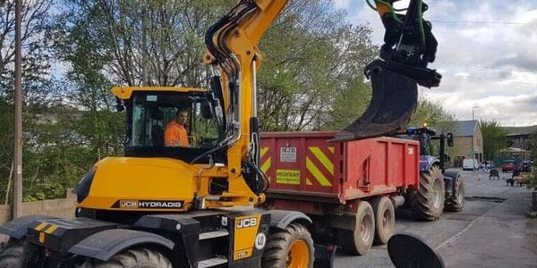 Yellow JCB excavator loading dirt into a red trailer on a road.