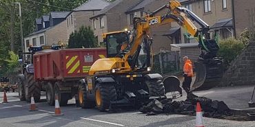 Construction workers using a JCB excavator to break up asphalt on a residential street.