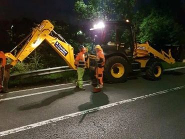 Two construction workers talk beside a JCB backhoe loader on a dimly lit road at night.