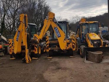 Three yellow backhoe loaders parked on a dirt surface with trees in the background.