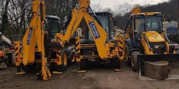Three yellow backhoe loaders parked on a dirt surface with trees in the background.