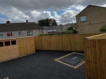 Backyard with black asphalt, wooden fencing, shed, and an in-ground trampoline under cloudy sky.