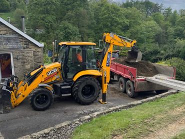 Yellow JCB backhoe loader dumping dirt into a red trailer beside a house.