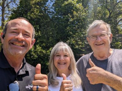 Smiling roofing contractor posing with happy customers outdoors, all giving thumbs up, with trees