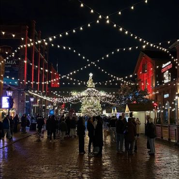 Crowded festive street at night with Christmas lights and a decorated tree.