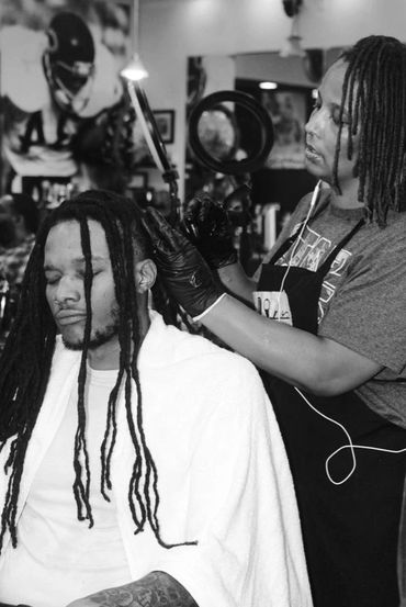 Barber carefully styling a man's dreadlocks in a busy salon.