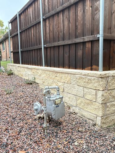Gas meter with pipes near a wooden fence and stone wall.