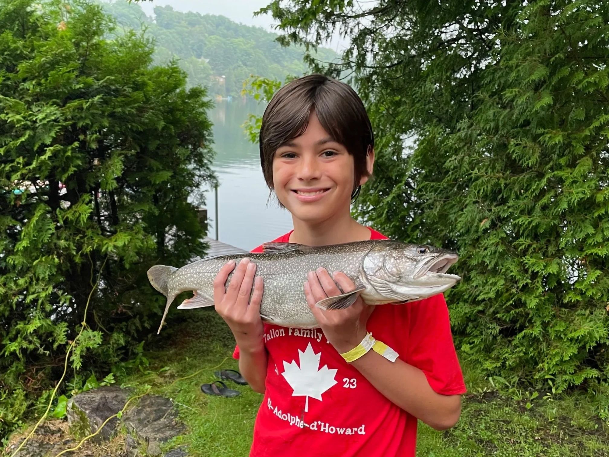 A boy in a red shirt proudly holds a large fish near a lakeside.