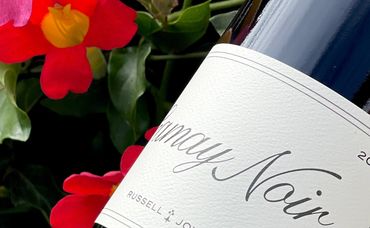 Close-up of a 2024 Gamay Noir wine bottle with vibrant red flowers and green leaves in the background.