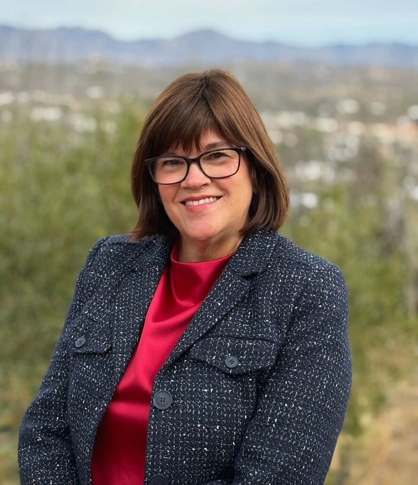 Smiling woman in glasses wearing a blue tweed jacket and red top outdoors.
