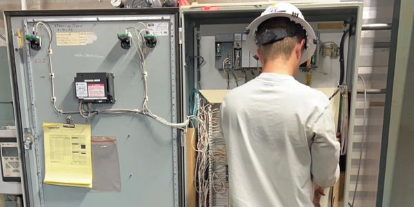 Technician wearing a hard hat working on an open electrical control panel.