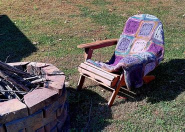 Wooden chair with a colorful blanket beside a brick fire pit on grass.