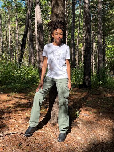 Young woman standing against a tree in a forest, wearing a white t-shirt and green cargo pants.
