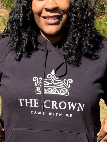 Woman smiling outdoors wearing a black hoodie with a crown design and text.