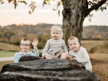 Three children sitting on rocks outdoors, one waving at the camera.