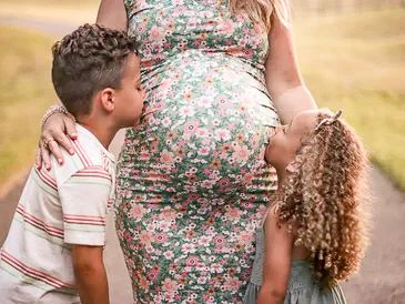 Two children kiss their pregnant mother's belly outdoors.