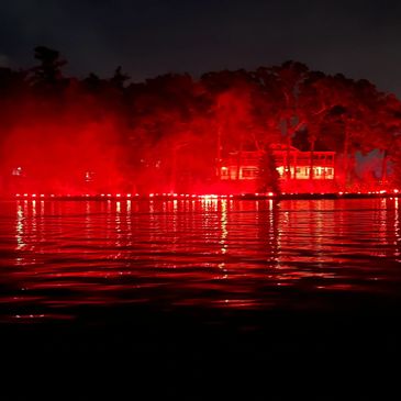 Red lights illuminate a waterfront scene at night with reflections on the water.