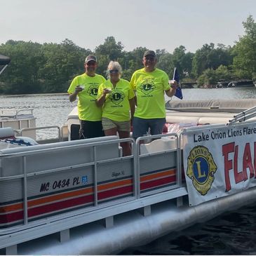 Three people in matching Lions Club shirts on a pontoon boat by a lake.