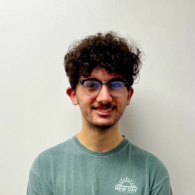 Young man with curly hair and glasses smiling against a plain wall.