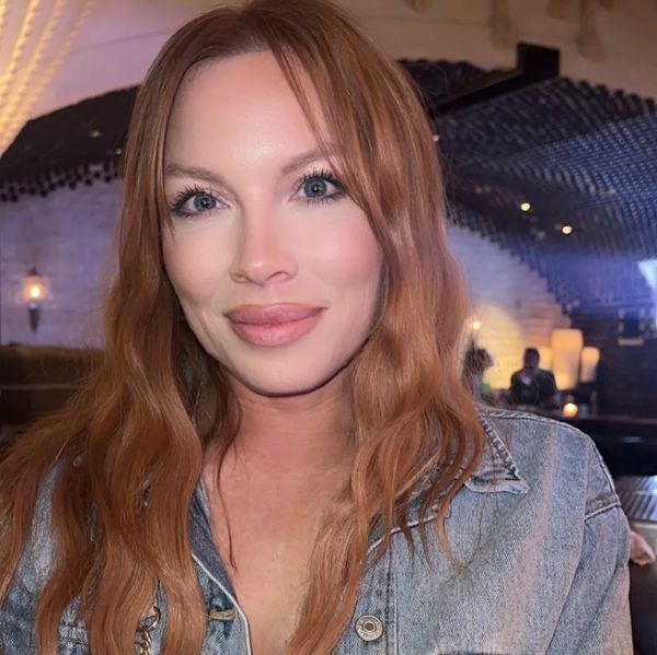 A woman with long red hair smiling indoors in a cozy, dimly lit setting.