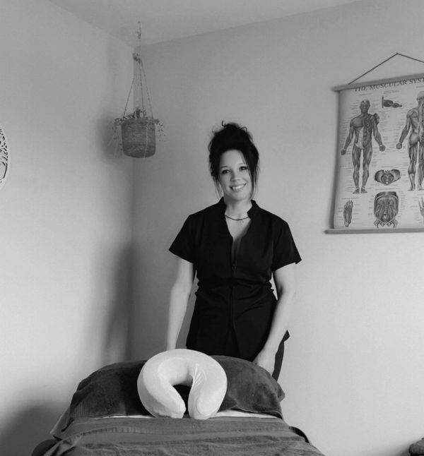 A smiling woman stands behind a massage table in a calm room.