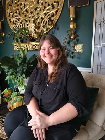 Woman smiling while seated indoors with plants and golden wall decor.