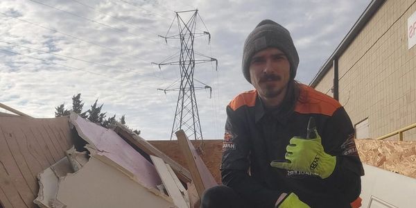 Worker in gloves poses on debris pile with cloudy sky and power lines in background.