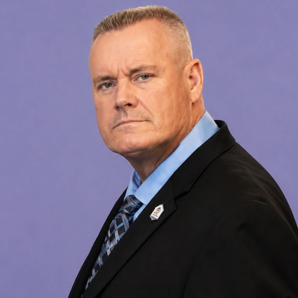 Serious man in a suit with a blue shirt and patterned tie against a purple background.