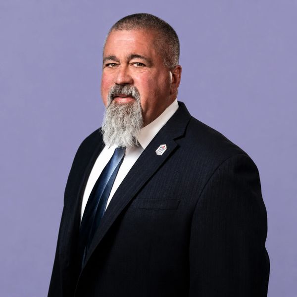 Man with a gray beard in a dark suit and tie against a purple background.