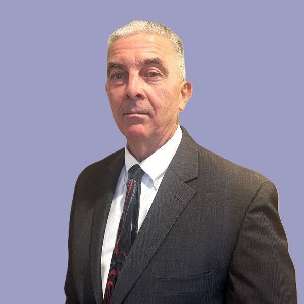 A confident older man in a dark suit and patterned tie against a plain background.