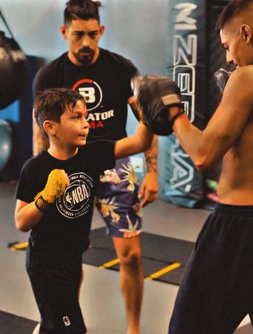 A young boy practices boxing punches with a trainer and coach in a gym.