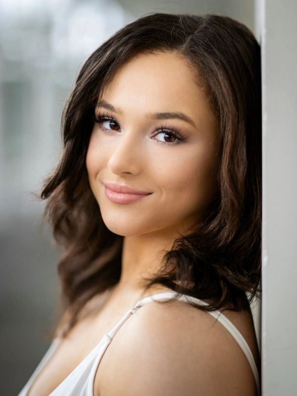 A young woman with dark hair smiling gently, leaning against a wall.