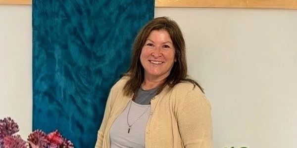 A smiling woman in a beige cardigan standing indoors near flowers.
