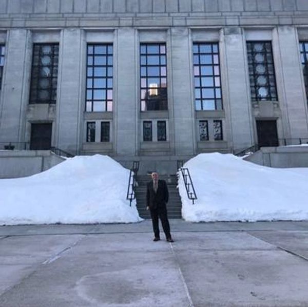 Man in suit stands in front of large building with snow piles on stairs.