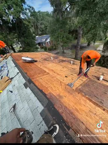 Two workers are repairing a roof, removing old shingles and preparing the surface.