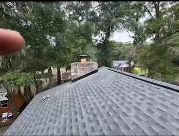 View of a shingled roof with a chimney and surrounding trees.