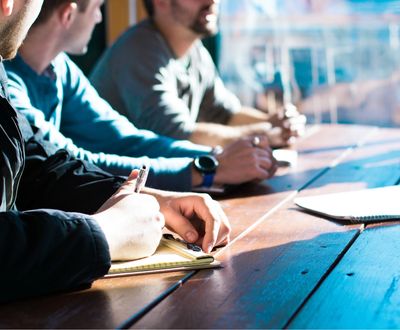 People taking notes during a meeting at a wooden table.