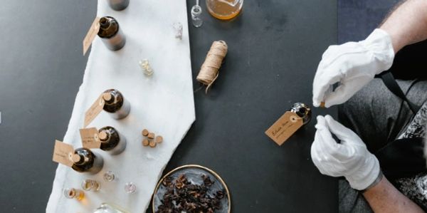 Person wearing white gloves handling glass bottles with essential oils and dried herbs on a table.