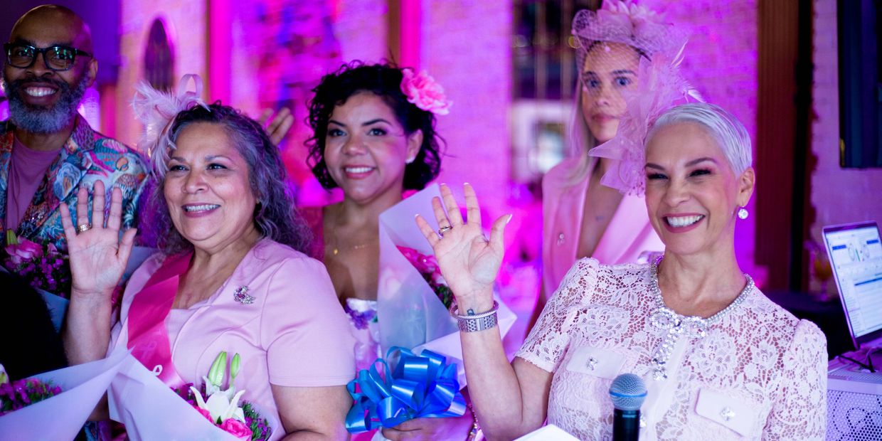 Group of women smiling and waving, dressed in pink with flowers and sashes.