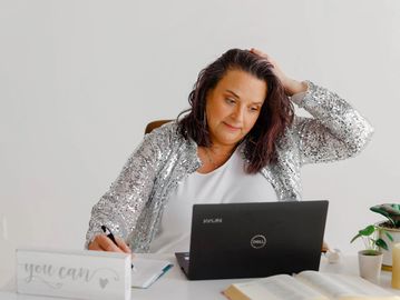 Woman in silver jacket working and writing while looking at laptop.