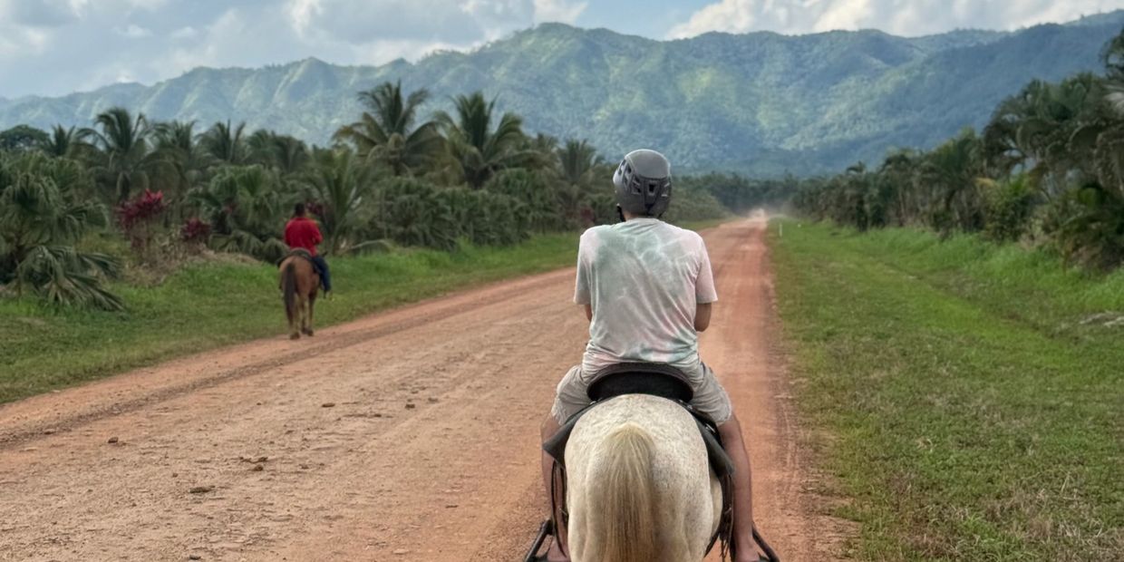 Two people horseback riding on a dirt road with mountains in the background.