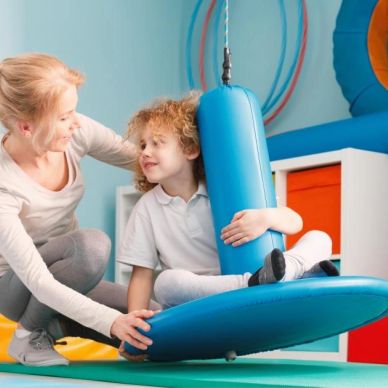 Therapist helping child on a blue swing seat in a colorful therapy room.