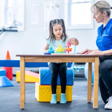 A young girl playing with colorful rings under therapist supervision in a gym.